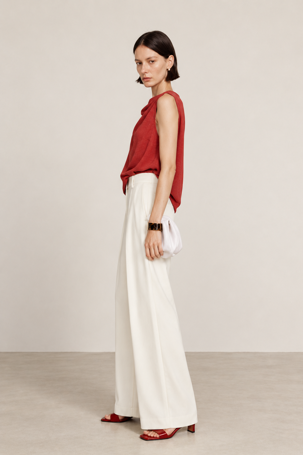 A woman stands in profile wearing a sleeveless red top, wide-leg white pants, red heeled sandals, and holding a white clutch bag against a plain beige studio background.