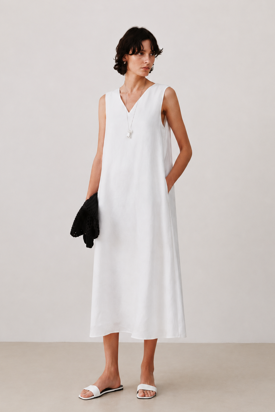 A woman modeling a sleeveless white midi dress with a V-neckline and a single decorative button, standing against a neutral light beige studio background.