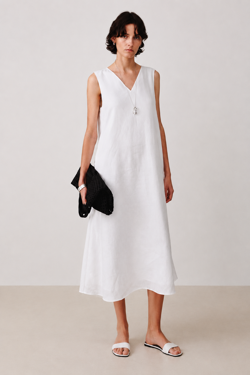 A woman modeling a sleeveless white linen midi dress with a V-neckline, holding a black clutch and wearing white slide sandals against a neutral studio backdrop.