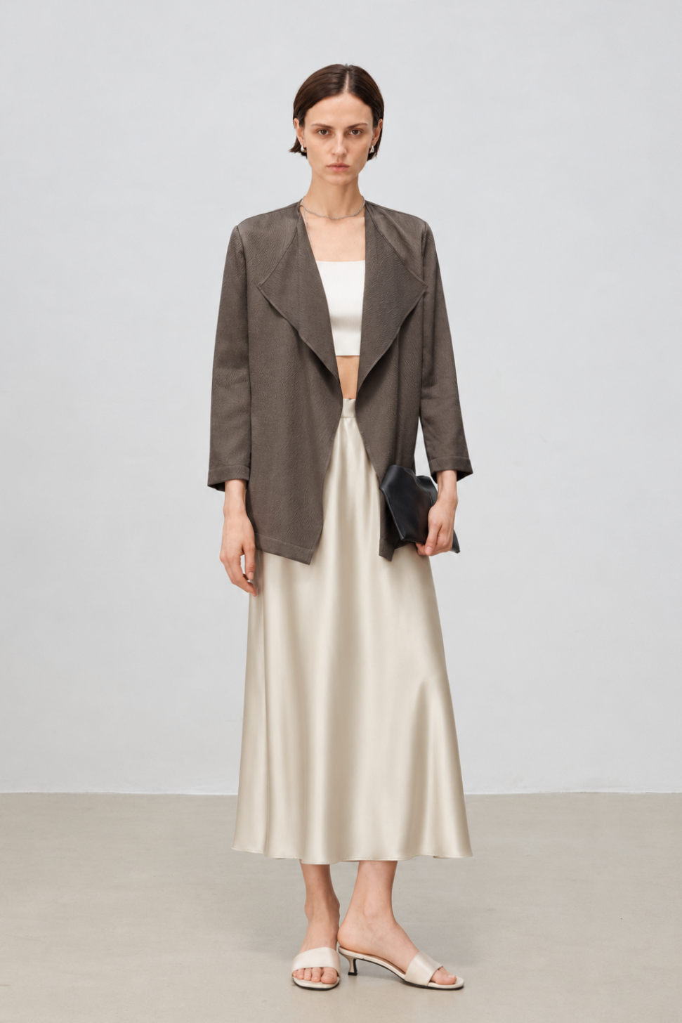 A model stands against a light gray studio background wearing a dark taupe open-front blouse over a white cropped top and a cream satin midi skirt, holding a small black clutch.