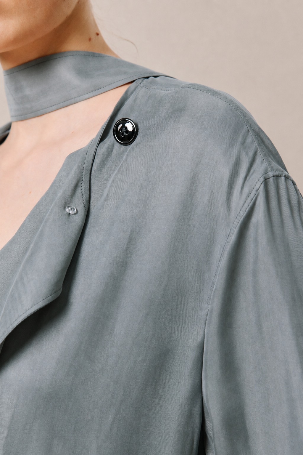 Close-up of a person wearing a slate gray blouse with a high mock neck, asymmetrical button detail, and a small cutout at the shoulder.