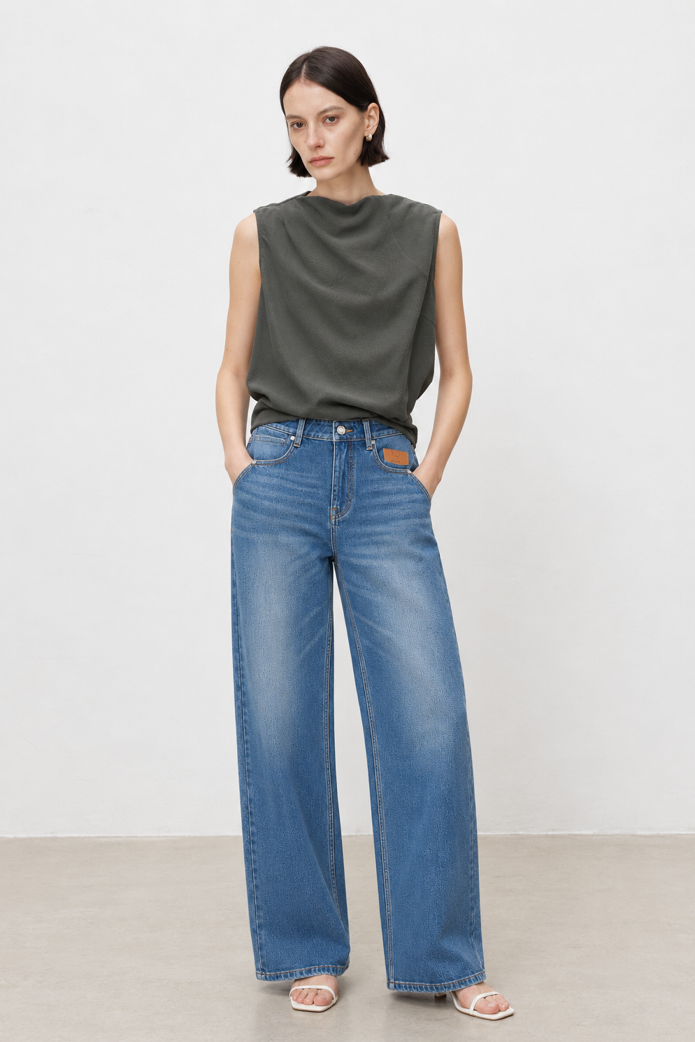 A woman modeling a dark olive green sleeveless top and wide-leg blue jeans against a plain light gray studio background.