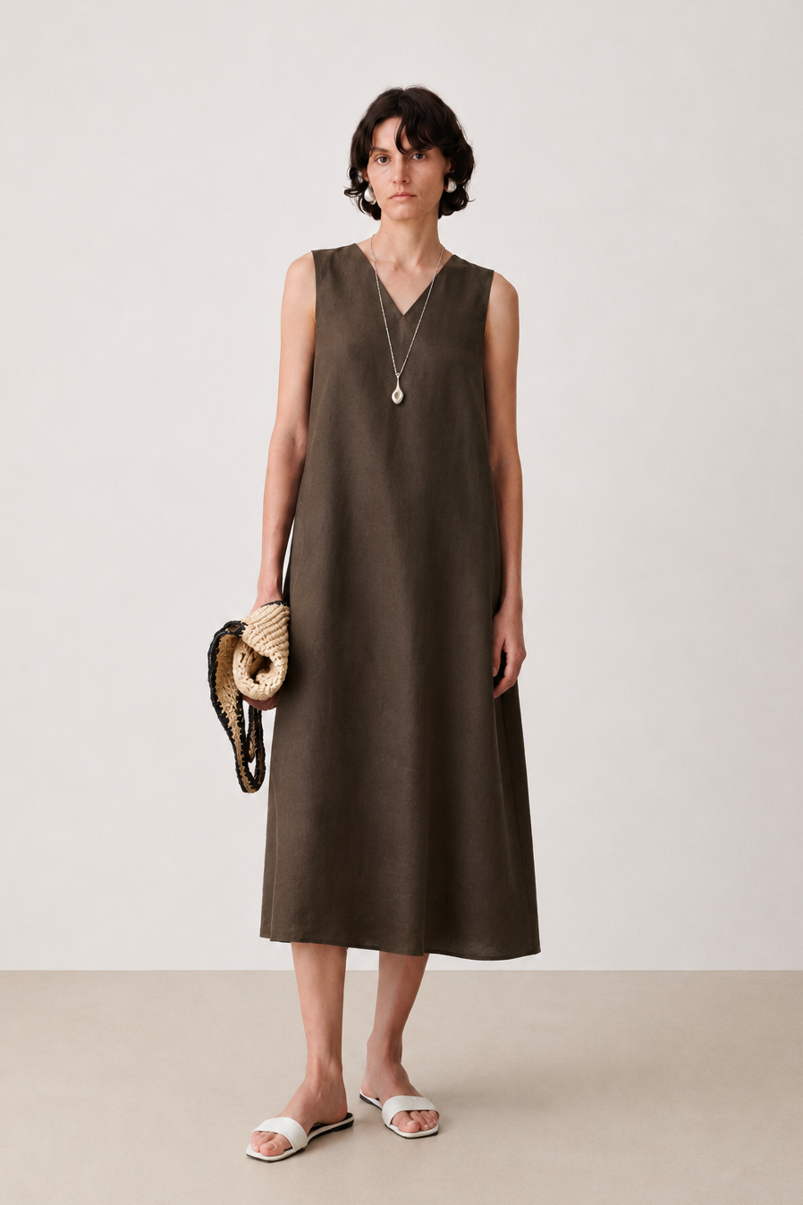 A woman stands in a minimalist studio wearing a sleeveless, V-neck midi dress in a dark brownish-green color, paired with white slide sandals, a long silver pendant necklace, and holding a woven straw clutch.
