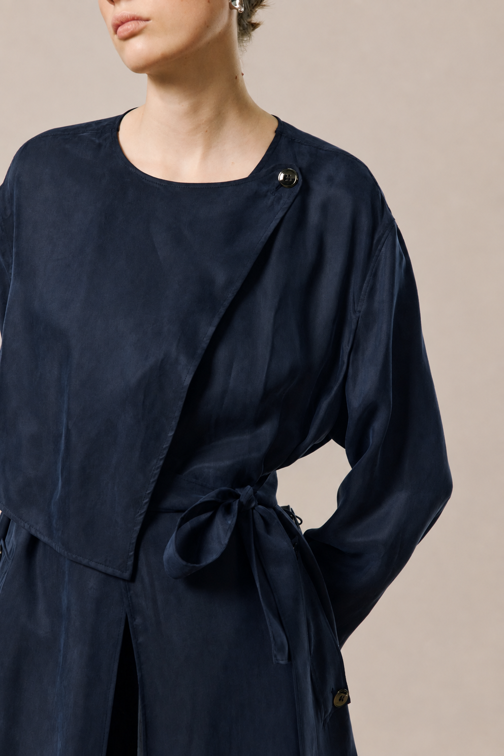 A close-up of a model wearing a navy blue wrap-style top or dress with a tie waist and small dark buttons near the shoulder, photographed against a plain warm beige studio background.