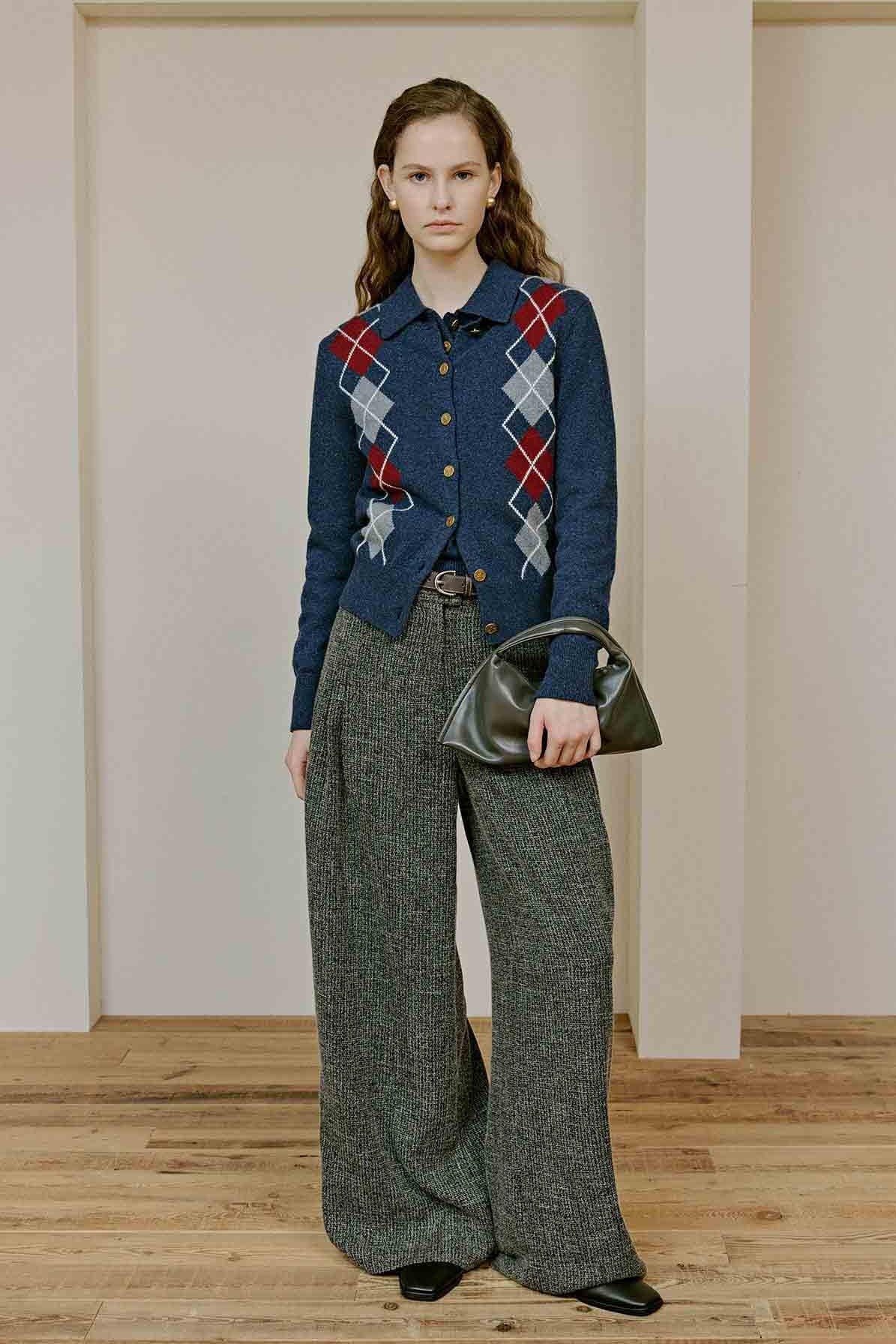Woman wearing a blue argyle sweater and gray wide-leg pants holding a green handbag indoors.