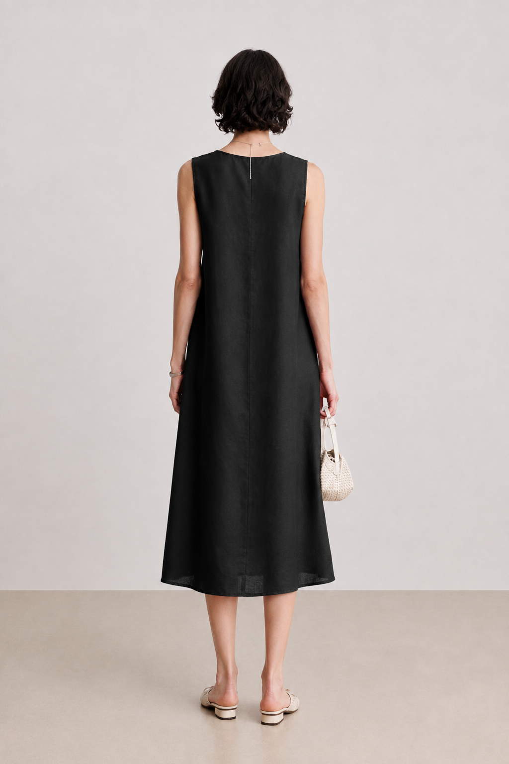A woman viewed from behind wearing a sleeveless black midi dress with a relaxed A-line silhouette. She is holding a small cream-colored handbag and wearing flat sandals, set against a minimalist studio background with a light gray wall and beige floor.