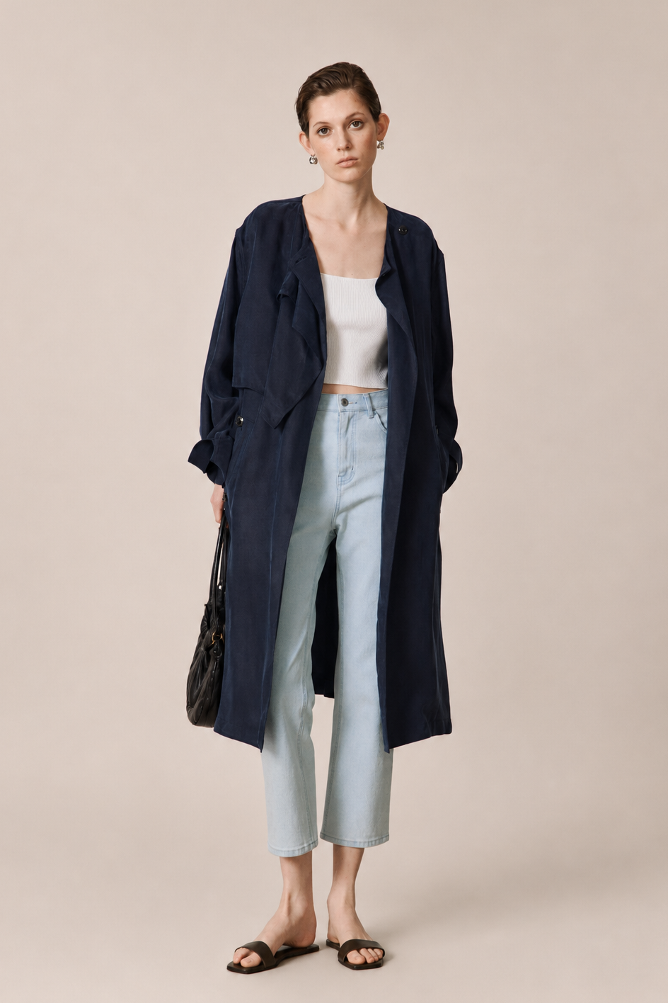 A model stands against a solid beige studio background wearing a long navy blue coat over a white crop top, light blue cropped jeans, and black slide sandals, while holding a black handbag.