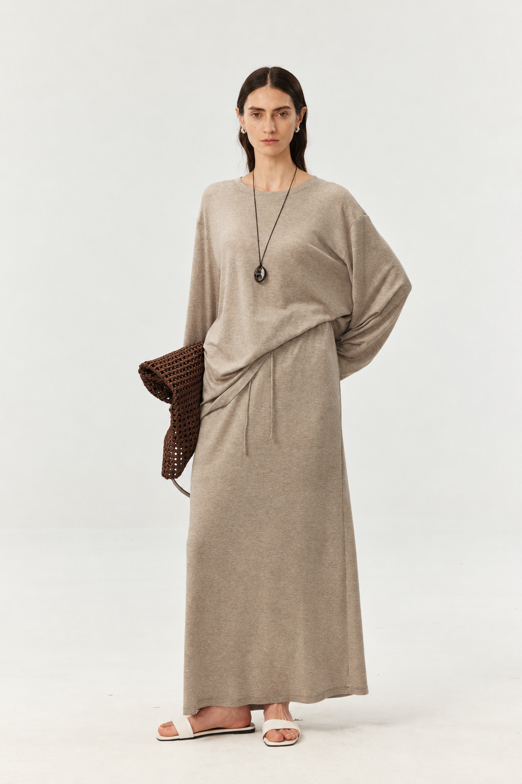A woman modeling a long-sleeved, draped maxi dress in a taupe-beige color, styled with a brown woven clutch and white slide sandals against a plain white studio background.
