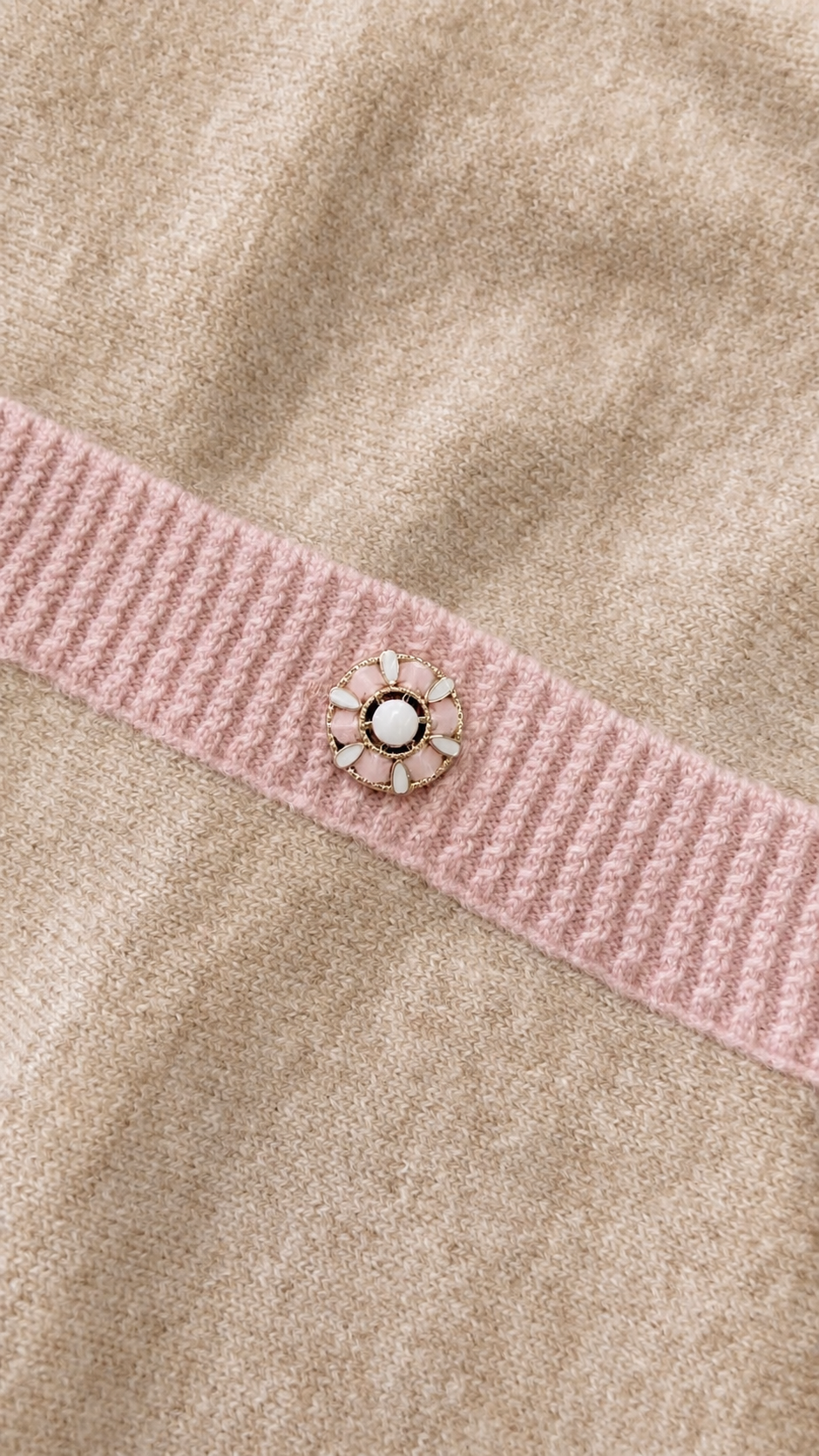 Close-up of a beige knit garment with a pink ribbed band and a small sunburst-style decorative embellishment featuring a white center and pale petals.