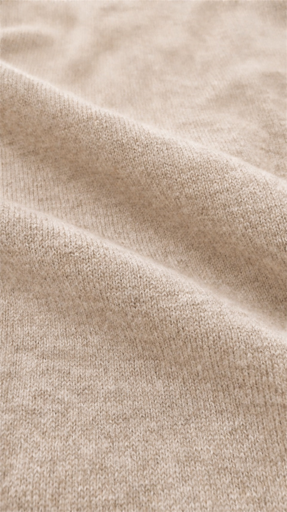 A close-up of soft, draped knit fabric in a warm light beige or oatmeal tone, showing the texture and weave detail.