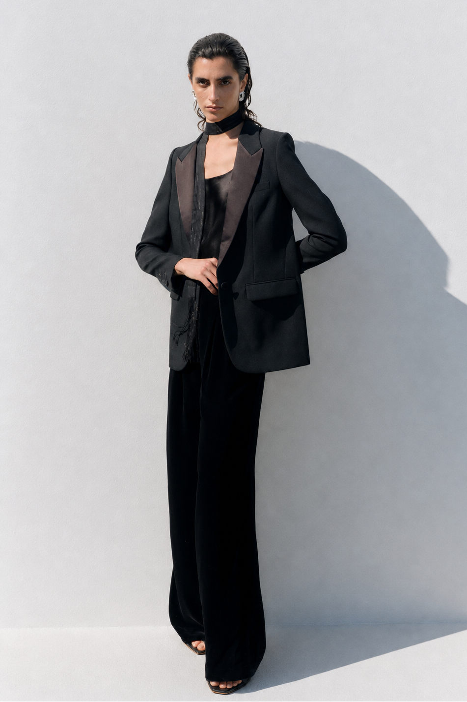 A model stands against a white wall wearing a black tailored blazer with satin lapels, a black choker, a black top, and wide-leg black trousers. Strong sunlight casts a sharp shadow on the wall.