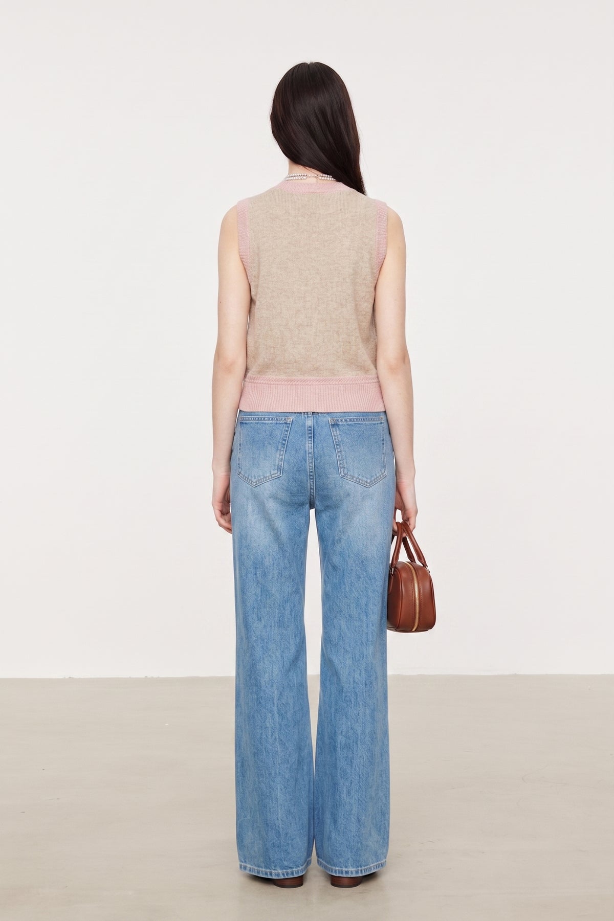 A woman is shown from behind wearing a beige knit sweater vest with a light pink ribbed hem, paired with wide-leg blue jeans and holding a small brown leather handbag against a clean, off-white studio background.