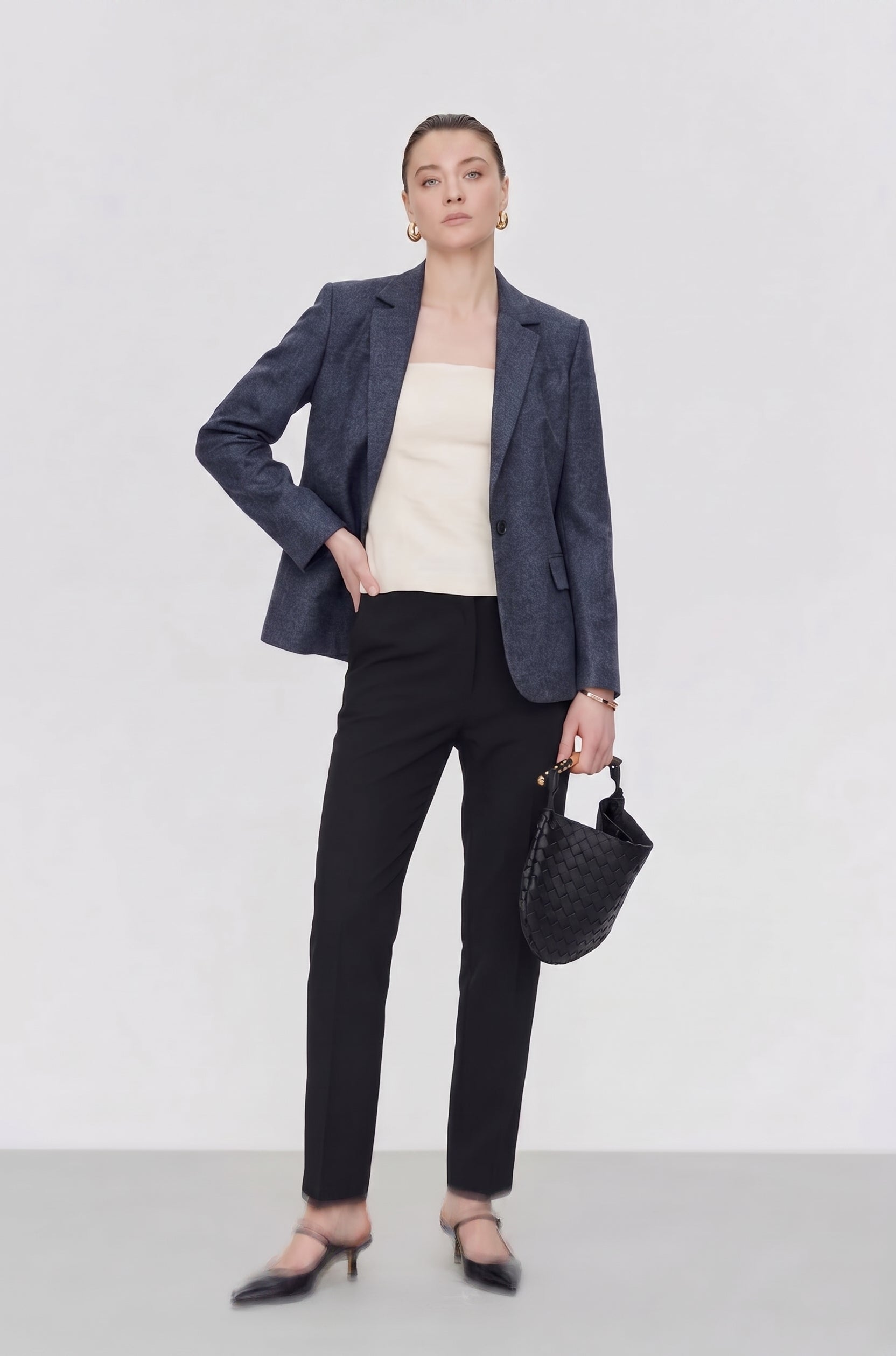 A woman models a dark blue textured blazer, black trousers, a cream top, and black pointed-toe mule heels, holding a black woven handbag against a light gray studio background.