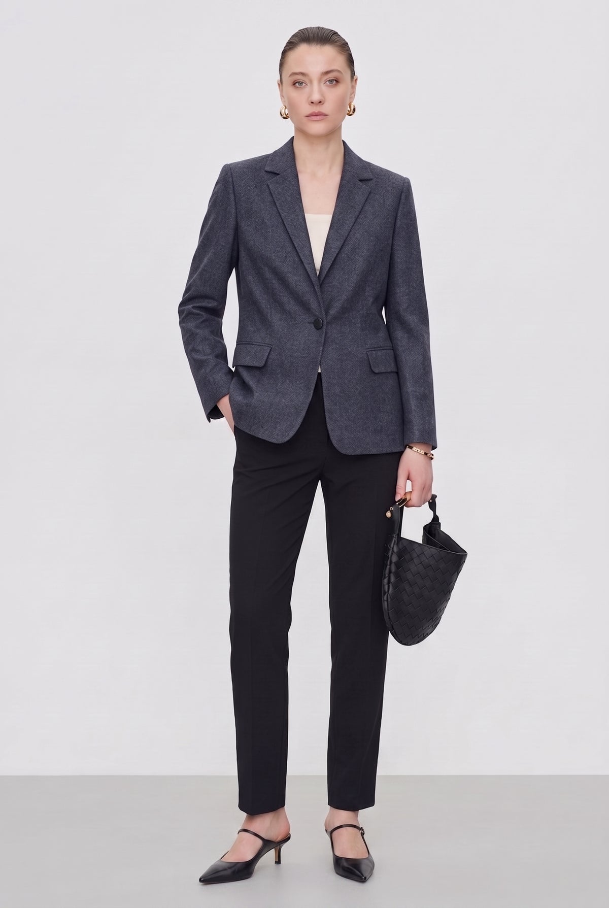 A model stands against a light gray studio background wearing a tailored dark gray blazer, black trousers, black pointed heels, and holding a black woven handbag.