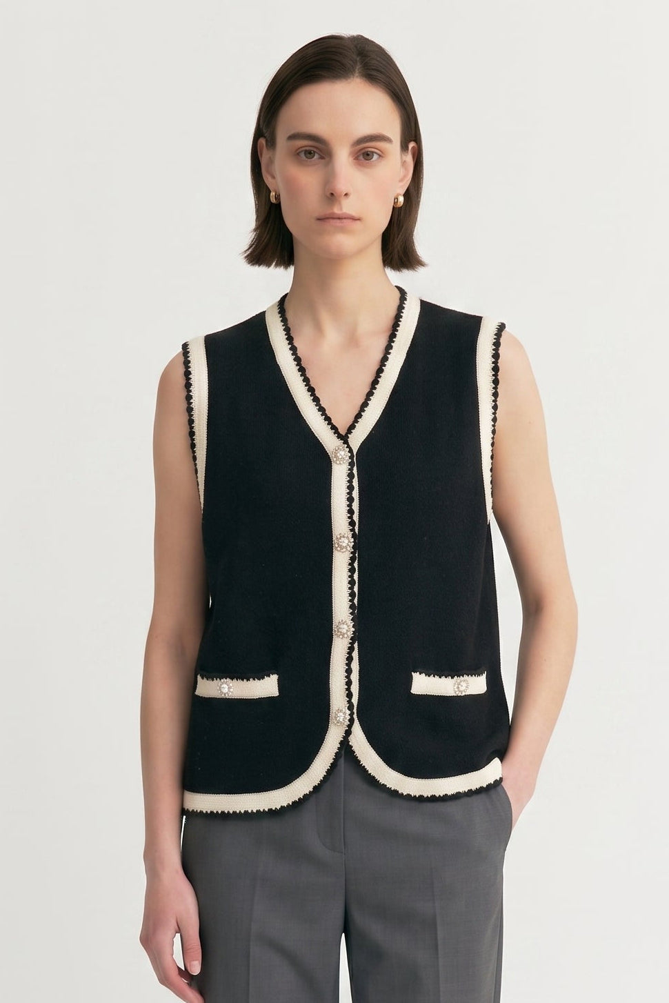 A model stands against a light gray studio background wearing a sleeveless black vest with cream trim, gray trousers, and black loafers.