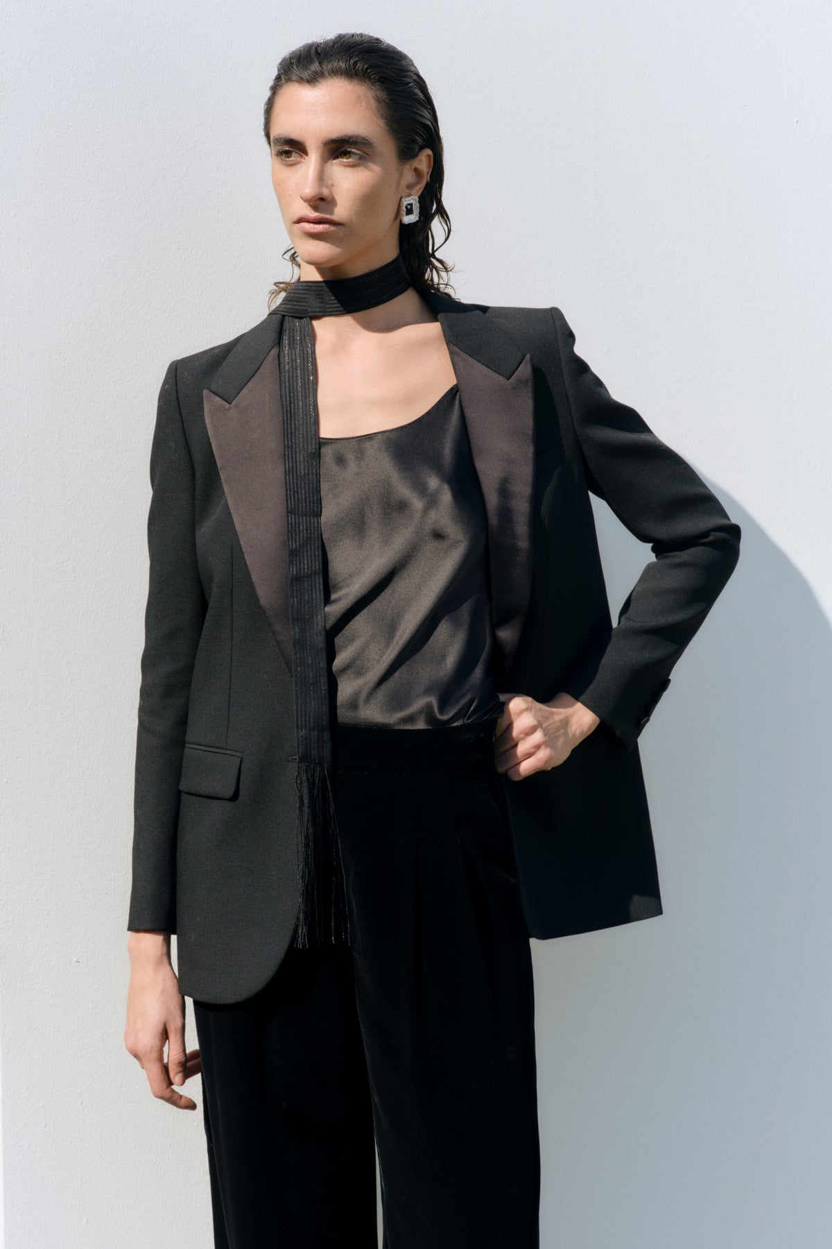 A model wearing a black tuxedo-style blazer with satin lapels, a black satin top, and black trousers, standing against a light gray wall.